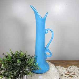 May include: A tall, light blue ceramic vase with a unique, elongated shape. It features a curved handle and a flared base. The vase is approximately 12 inches tall and is displayed on a wooden surface with a decorative lace doily.