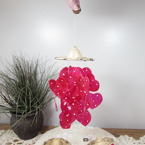 May include: A hanging mobile featuring a small woven hat above a cluster of red heart-shaped decorations with white polka dots. The mobile is suspended by a thin string. A potted plant is visible in the background.