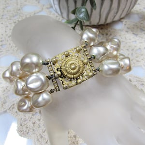 May include: Vintage pearl bracelet featuring large, irregular faux pearls. The bracelet has a decorative gold-tone clasp with an ornate, embossed design. The pearls are a creamy off-white color, and the bracelet is displayed on a mannequin hand.