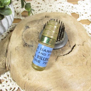 May include: A cylindrical brass thimble featuring a light blue paper label with dark blue text that reads THE BARRE, R AND FE, A MILLING, ONE 799. The top of the thimble has a textured, dimpled surface, while the base is smooth and metallic.