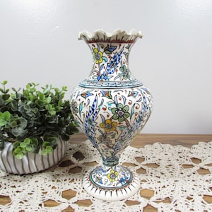 May include: A ceramic vase with a ruffled rim and a pedestal base, featuring a white background decorated with intricate, multi-colored floral patterns in shades of blue, green, yellow, and brown, covering the entire exterior surface.
