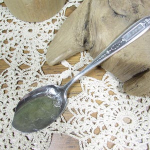 May include: A silver-toned metal spoon featuring an elongated handle with intricate embossed scrollwork and a rectangular decorative panel. The bowl of the spoon is smooth and oval-shaped, showing a reflective surface with minor surface scratches.