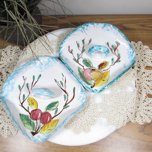 May include: Two white ceramic dishes with a blue rim and a fruit design. Each dish has a raised center with a small handle. The fruit design includes red cherries, green leaves, and yellow and orange fruit.
