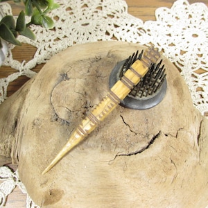 May include: A light tan, tapered wooden tool with dark brown carved bands and a pointed tip, resting on a circular black metal flower frog with numerous upright metal pins. The tool features a textured, carved surface with repeating geometric patterns.