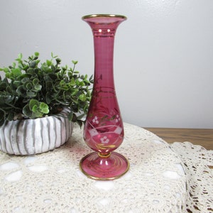 May include: A tall, ruby-colored glass vase with a flared top and a round base. The vase has gold trim along the top and base, and delicate gold and white floral designs. The vase is approximately 10 inches tall.