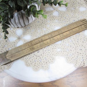 May include: A vintage wooden folding ruler, measuring up to 24 inches. The ruler is light brown with dark markings and metal hinges. It is resting on a white surface with a cream-colored lace doily.