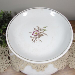 May include: White ceramic bowl with a floral design in the center. The flower has purple and yellow petals with brown and green leaves. The rim of the bowl has a gold trim. The bowl is sitting on a cream-colored lace doily.