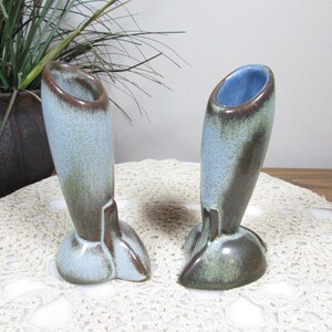 May include: Two ceramic bud vases with a unique shape. The vases have a light blue exterior with brown accents and a blue interior. The base is rounded with a fin-like detail. The vases are displayed on a white lace doily.