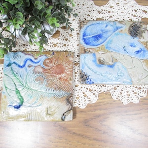 May include: Two square ceramic coasters with nature-inspired designs. One features a green leaf and blue swirls, the other has blue leaf and abstract patterns. The coasters are in shades of beige, brown, and blue.