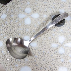 May include: A vintage silver ladle with a round bowl and long handle, resting on a cream-colored, crocheted doily. The handle features ornate floral detailing. The ladle is likely used for serving soups or sauces.
