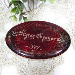 May include: Oval, ruby-red glass object with etched text reading "Margee Keener - 1907." The glass has a textured appearance with gold accents. The object is set on a cream-colored, lace-like surface.