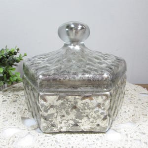 May include: A vintage-style, square glass jar with a lid. The jar has a textured, weathered appearance with a silver finish. The lid features a rounded knob. The jar sits on a white lace doily.