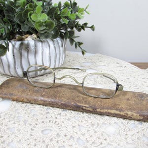 May include: Antique eyeglasses with a gold-toned metal frame and oval lenses rest on a worn, brown leather case. A decorative white and gray striped ceramic pot with greenery sits in the background, on a white lace tablecloth.