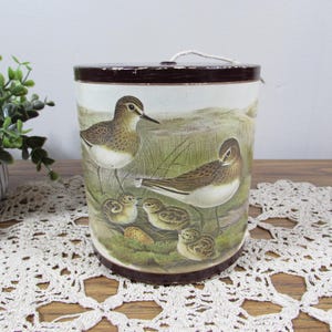 May include: A cylindrical tin container with a dark brown lid and a white body featuring a detailed illustration of two adult sandpiper birds and three chicks in a grassy, natural landscape. The container has a small white string loop on top.