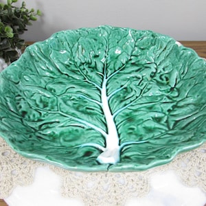 May include: A green ceramic cabbage leaf-shaped serving dish. The dish features a detailed vein pattern and a white stem. The edges of the dish are scalloped. This decorative piece is suitable for serving food.