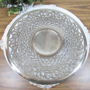 May include: A silver-colored, decorative bowl with an intricate, openwork design. The bowl features a central, solid base and a detailed, floral-inspired pattern around the rim. It rests on a white, lacy doily, suggesting a vintage or antique aesthetic.