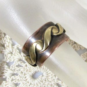 May include: A wide band ring featuring a dark copper-toned base with a polished gold-toned metal overlay. The overlay is shaped into a continuous, repeating wave or scroll pattern that wraps around the circumference of the dark metal band.