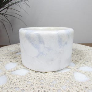 May include: A cylindrical, white marble container with subtle gray veining. The small, open-topped vessel sits on a cream-colored, crocheted doily. The background includes a dark vase with greenery.