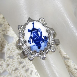 May include: A silver-toned ring featuring an oval, white ceramic centerpiece with a blue windmill design. The ring has an ornate, openwork setting with decorative details. The ring is a piece of jewelry.