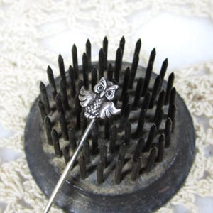 May include: A silver-toned owl-shaped hat pin is centered on a dark, circular flower frog. The pin features an owl with detailed wings and eyes. The flower frog has many dark, sharp pins. The background is a cream-colored, open-weave fabric.
