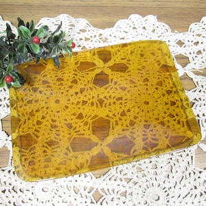 May include: A rectangular amber-colored glass tray featuring an embedded yellow crochet doily pattern. The tray is accented with a small decorative sprig of green leaves and two red berries positioned on the top left corner of the glass surface.