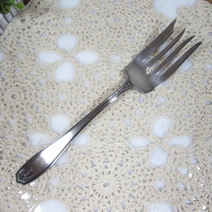 May include: A silver serving fork with four tines, resting on a cream-colored, crocheted doily. The fork has a decorative handle with an ornate design. The doily features a floral pattern.