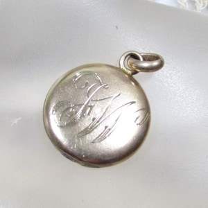 May include: A small, round, silver-toned pendant with a loop for a chain. The pendant features engraved cursive initials. The pendant is a vintage piece, likely a locket or charm, with a simple, elegant design.