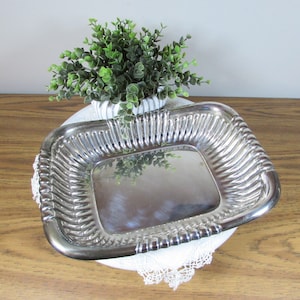 May include: A rectangular silver-colored tray with a ribbed design on the sides. The tray is placed on a white lace doily and a wooden surface. A small white pot with green foliage is behind the tray.