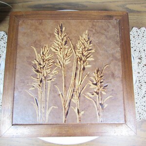 May include: Square wooden decorative tray with a wheat design. The wheat stalks are in a light brown color, set against a textured, brown background. The tray has a dark brown wooden frame.