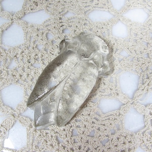 May include: A clear, glass owl figurine with a textured surface. The owl is lying on a cream-colored, crocheted doily. The owl's wings are slightly spread, and the details of the owl's face are visible.