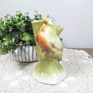 May include: A ceramic bird figurine perched on a tree trunk, painted with blue, orange, and yellow hues. The bird is set against a backdrop of a green plant in a decorative pot and a cream-colored lace doily.