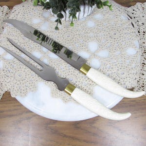 May include: A carving knife and fork set with ivory-colored handles and gold-colored accents. The knife has a long, serrated blade, and the fork has two tines. The set is displayed on a white plate with a decorative lace doily.