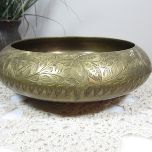 May include: A brass-colored, round bowl with intricate leaf and floral patterns etched around the exterior. The bowl sits on a white, lace-like doily. A dark brown pot with greenery is visible in the background.