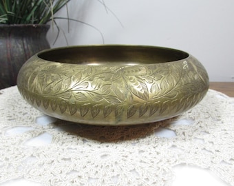 May include: A brass-coloured, round bowl with intricate leaf and floral patterns etched around the exterior. The bowl sits on a white, lace-like doily. A dark brown pot with greenery is visible in the background.