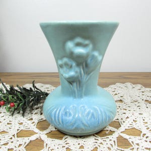 May include: A light blue ceramic vase with a flared rim and a rounded base. The exterior features a raised relief design of three tulip-like flowers on the neck and a decorative pattern of leaves and stems encircling the lower body.