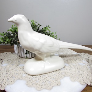 May include: A white ceramic bird figurine with a long tail and a smooth, glossy finish. The bird is perched on a rounded base. A small potted plant is in the background. The figurine is displayed on a decorative lace doily.