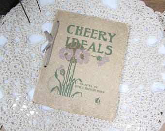 May include: A vintage book titled "Cheery Ideals" with a floral illustration on the cover. The cover is a light tan color with green and purple floral accents. The book is tied with a ribbon and rests on a cream-colored, crocheted doily.
