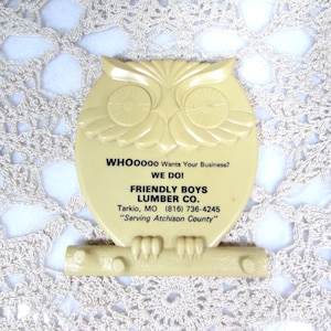 May include: A cream-colored owl-shaped sign with the text "WHOOOOO Wants Your Business? WE DOI FRIENDLY BOYS LUMBER CO." The sign is perched on a branch-like base and is set against a white lace background.