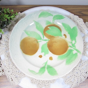 May include: A white ceramic plate with a decorative rim and a painted design of oranges and green leaves. The plate is set on a white surface with a beige lace doily.