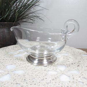 May include: Clear glass gravy boat with a silver-colored base and handle. The gravy boat is sitting on a white lace doily. A potted plant is in the background.