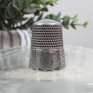 May include: A silver thimble with a perforated top and engraved design. The thimble has a detailed pattern around the base, featuring a central emblem. The thimble is a sewing tool, and is displayed on a clear glass surface.