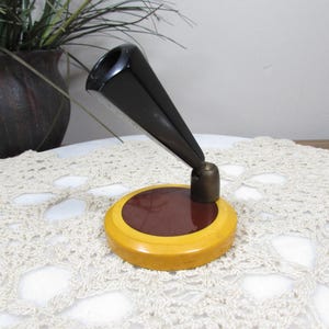 May include: A vintage pen holder with a black conical pen rest and a round base. The base has a dark red center and a yellow outer ring. The pen holder is set on a white lace doily.