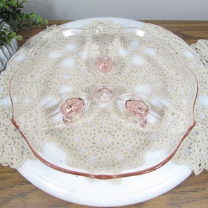 May include: A clear glass serving dish with a pink rim and three pink rose-shaped accents. The dish is placed on a white plate and a beige crocheted doily. The dish has a scalloped edge.