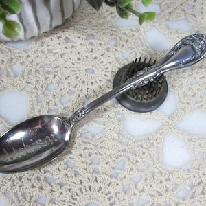 May include: A silver spoon with an ornate floral design on the handle and bowl. The word "Johnson" is engraved on the spoon's bowl. The spoon rests on a floral frog, with a cream-colored crocheted doily in the background.