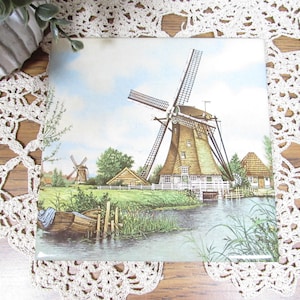 May include: Square ceramic tile featuring a picturesque scene of a Dutch windmill on a sunny day. The image shows a wooden boat, water, and lush greenery. The windmill has large blades and a thatched roof, evoking a sense of European charm.
