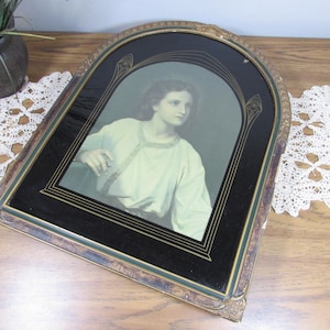 May include: A vintage framed picture of a young person in a white robe. The frame is gold with a black background and a green inner border.