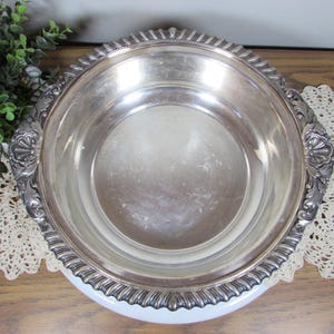 May include: A silver-colored, round serving bowl with a decorative, ridged rim and ornate handles. The bowl sits on a white base and is displayed on a wooden surface with a lace doily and greenery in the background. A vintage piece.