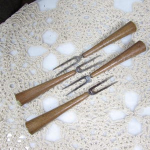 May include: Four vintage serving forks with wooden handles and metal tines are arranged on a cream-colored, open-weave doily. The forks have a classic design, with a warm brown hue to the wooden handles.