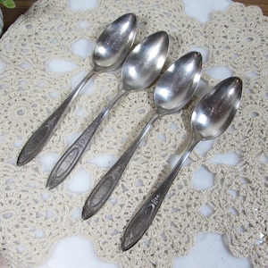 May include: A set of four silver-toned metal spoons arranged diagonally. Each spoon features a polished oval bowl and a slender handle decorated with an embossed floral pattern near the base and a smooth, elongated oval center section.