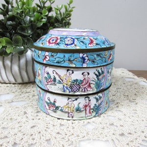 May include: A three-tiered, antique-style trinket box with a turquoise background and floral and figural painted designs. The box has a brass rim and a lid with a floral pattern. The box is sitting on a cream-colored lace doily.